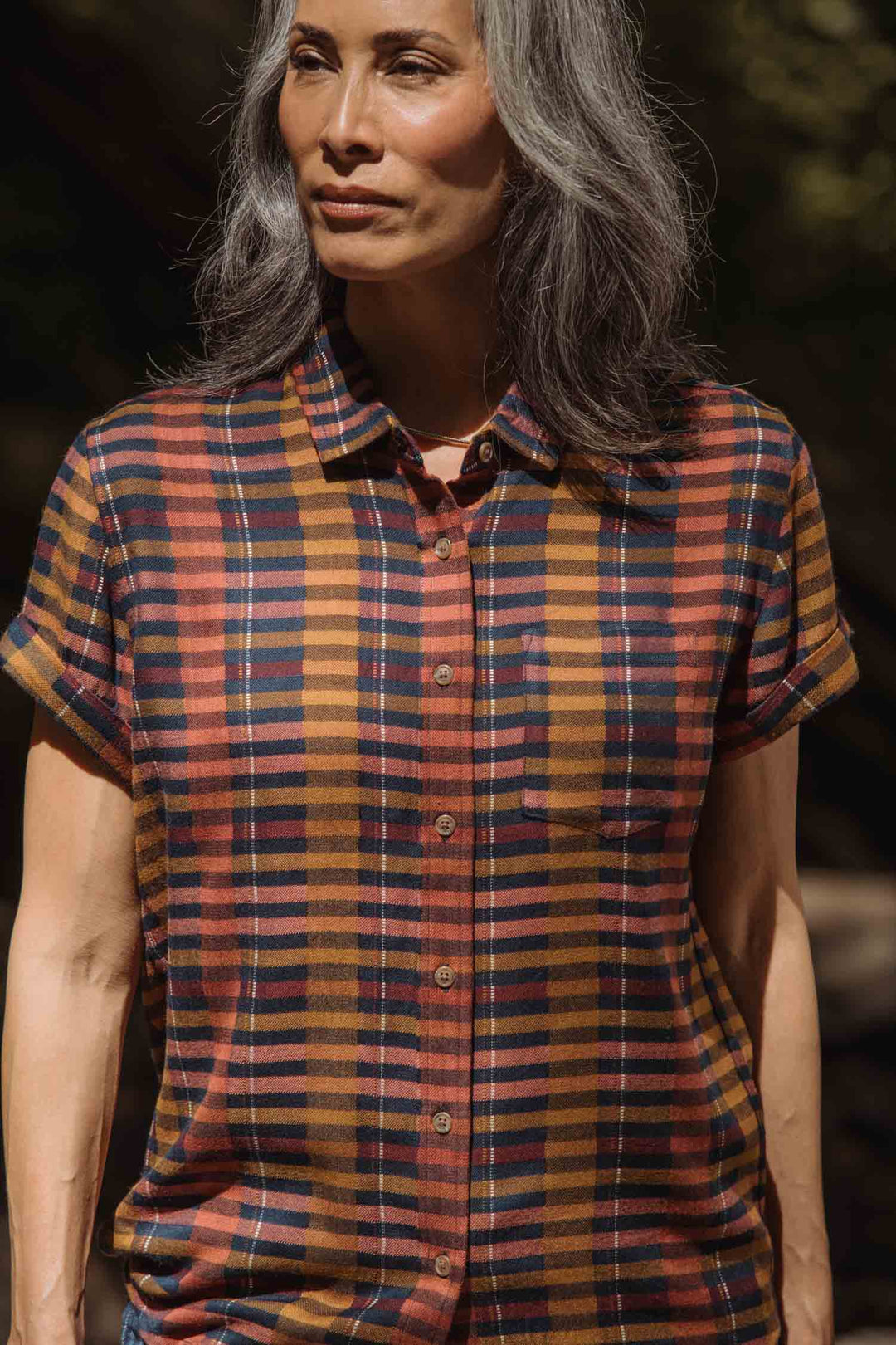 A person with long gray hair wears the Bridge & Burn Bea Shirt in Alpenglow Stripe, a short-sleeve button down made from an eco-friendly cotton alternative, standing outdoors in natural light.