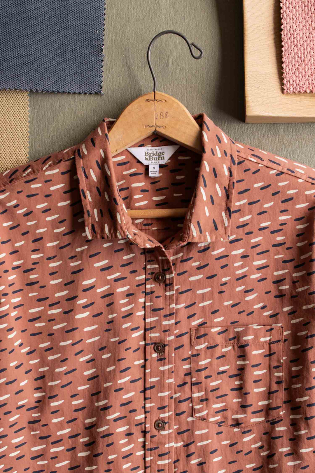 The Bea Shirt / Desert Rain by Bridge & Burn, a brown short-sleeve button-down with white and black dash patterns, hangs on a wooden hanger with textured fabric swatches in the background.