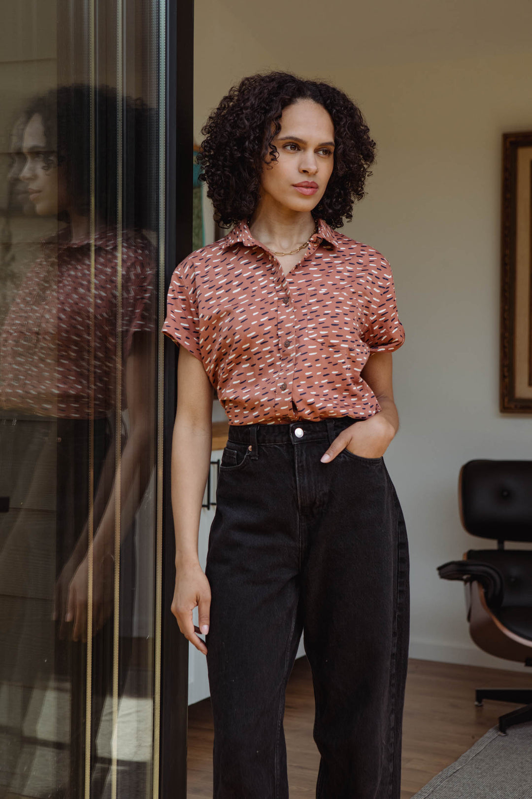 A woman with curly hair stands indoors by a glass door, wearing the Bridge & Burn Bea Shirt in Desert Rain and black high-waisted jeans, one hand in her pocket and a neutral expression. Her reflection appears in the glass.
