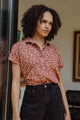 A woman with curly hair wears the Bridge & Burn Bea Shirt in Desert Rain and black pants, standing indoors by a glass door, gazing thoughtfully into the distance.