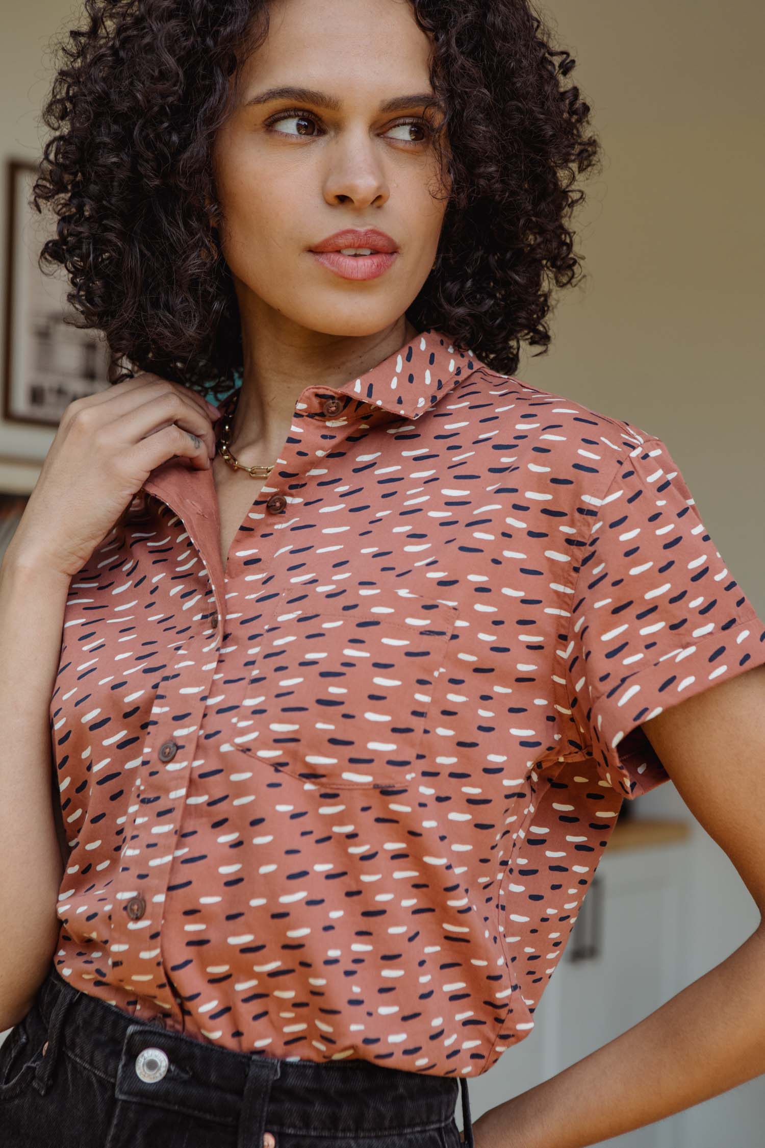 A woman with curly hair stands indoors, looking to the side, wearing the Bridge & Burn Bea Shirt / Desert Rain—a short-sleeve button-down patterned in brown, black, and white.