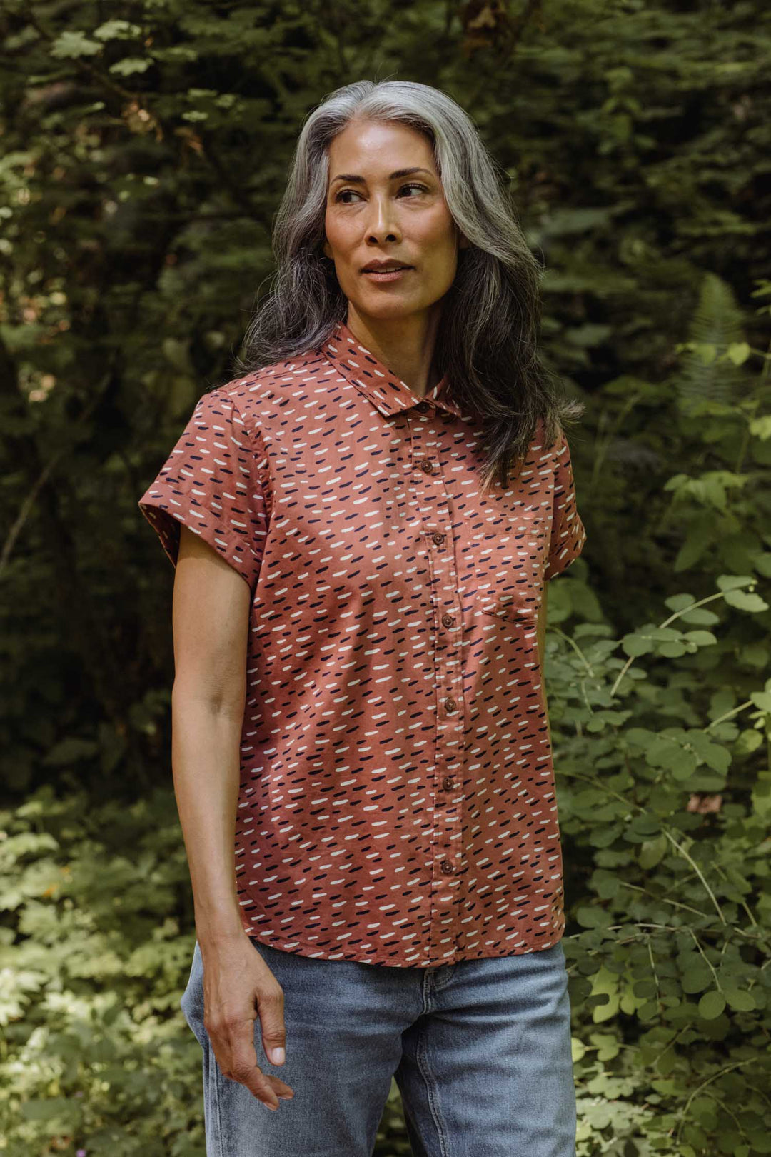 A woman with long gray hair stands outdoors among lush green plants and trees, wearing the Bridge & Burn Bea Shirt in Desert Rain and jeans, looking slightly to the side.