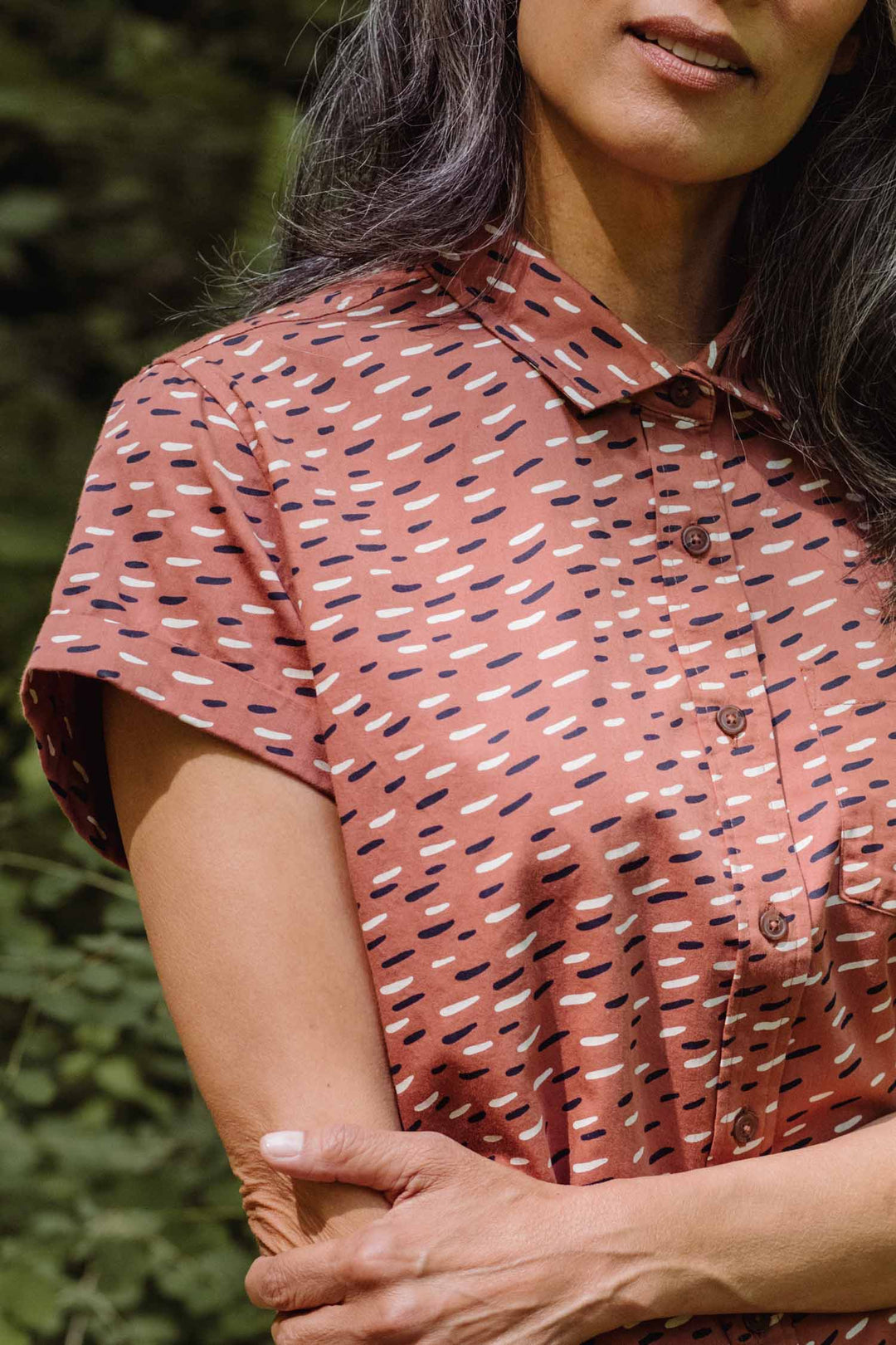 A woman with long, wavy gray hair stands outdoors, arms crossed before lush foliage, wearing the Bridge & Burn Bea Shirt / Desert Rain—a rust short-sleeve button-down patterned with scattered dashes.