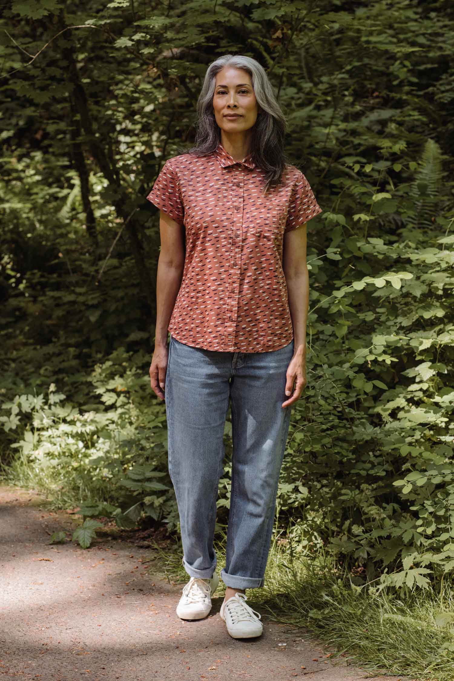 A woman with long gray hair stands on a leafy forest path, smiling gently. She wears the Bridge & Burn Bea Shirt in Desert Rain, paired with light blue jeans and white sneakers.