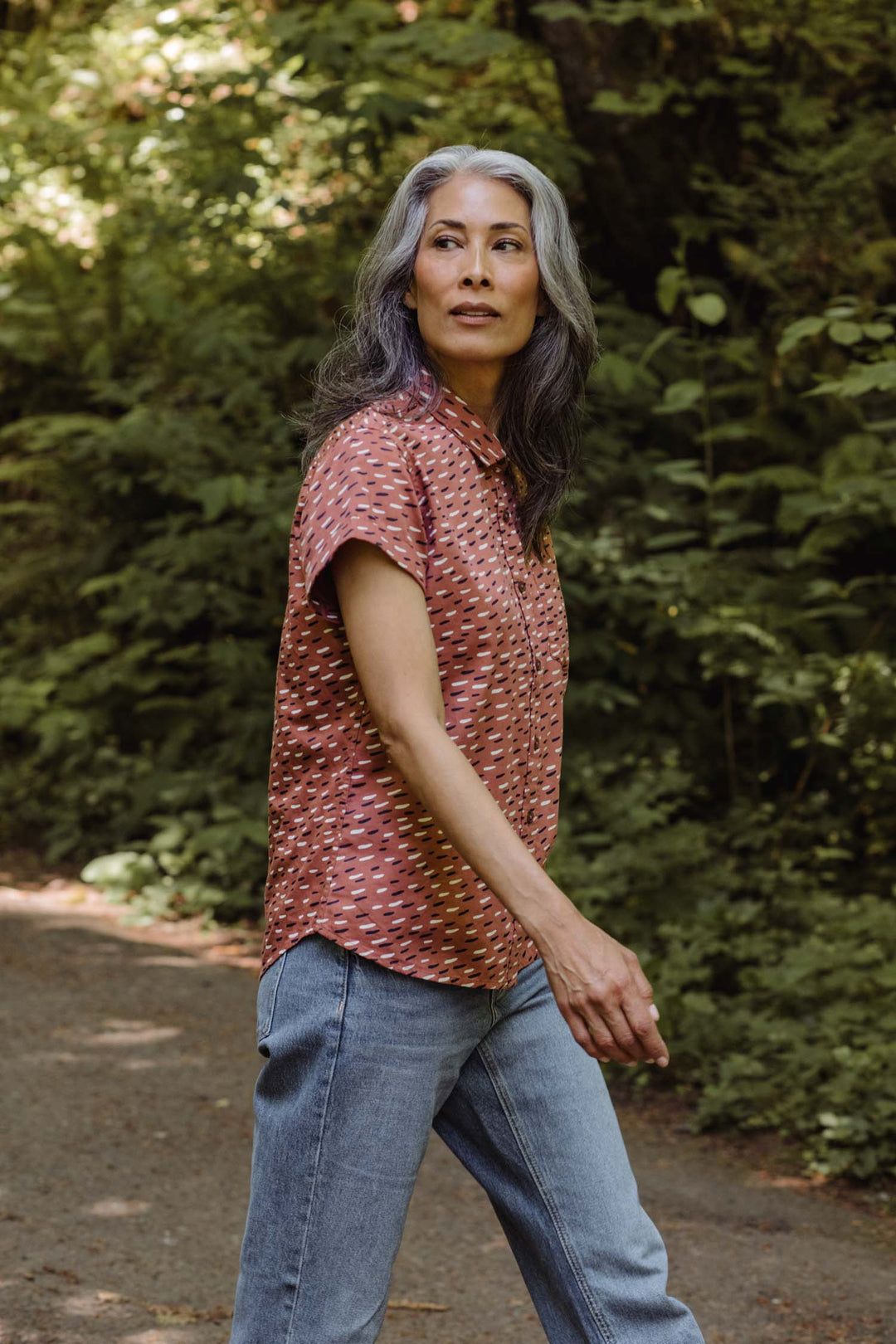 A woman with long gray hair walks outdoors on a path surrounded by green foliage, wearing the Bridge & Burn Bea Shirt in Desert Rain and blue jeans, looking to the side.