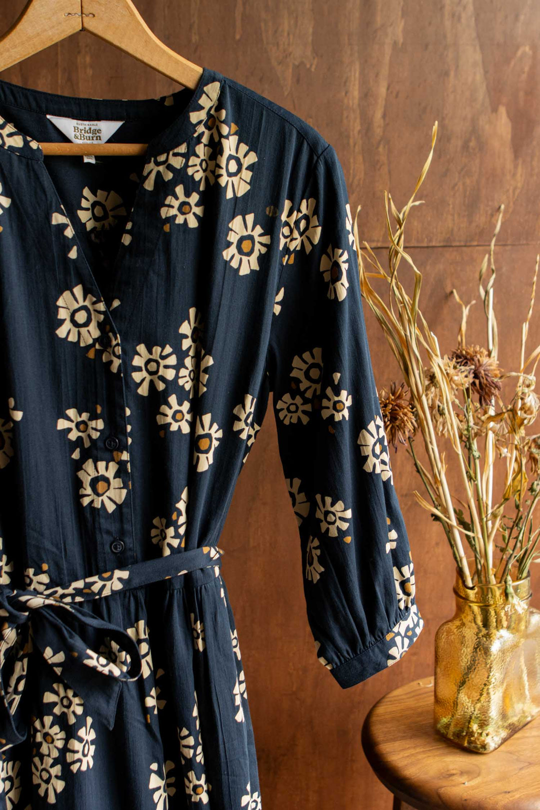 The Bridge & Burn Fiona Dress / Night Garden, featuring a navy organic cotton fabric with cream florals and gathered sleeves, hangs on a wooden hanger beside a gold vase of dried branches against a brown wooden backdrop.