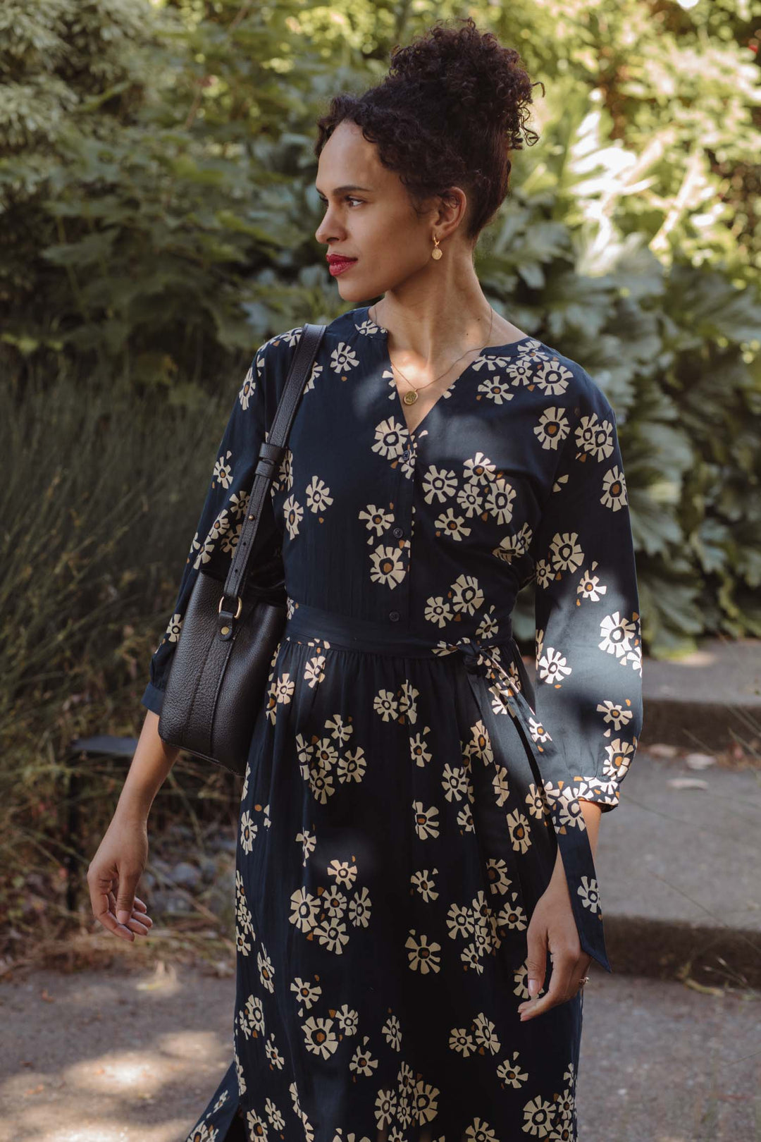 A woman with curly hair in a bun wears the Bridge & Burn Fiona Dress in Night Garden, a navy cream floral dress, paired with gold earrings and a black shoulder bag as she walks outdoors on a sunny day surrounded by greenery.