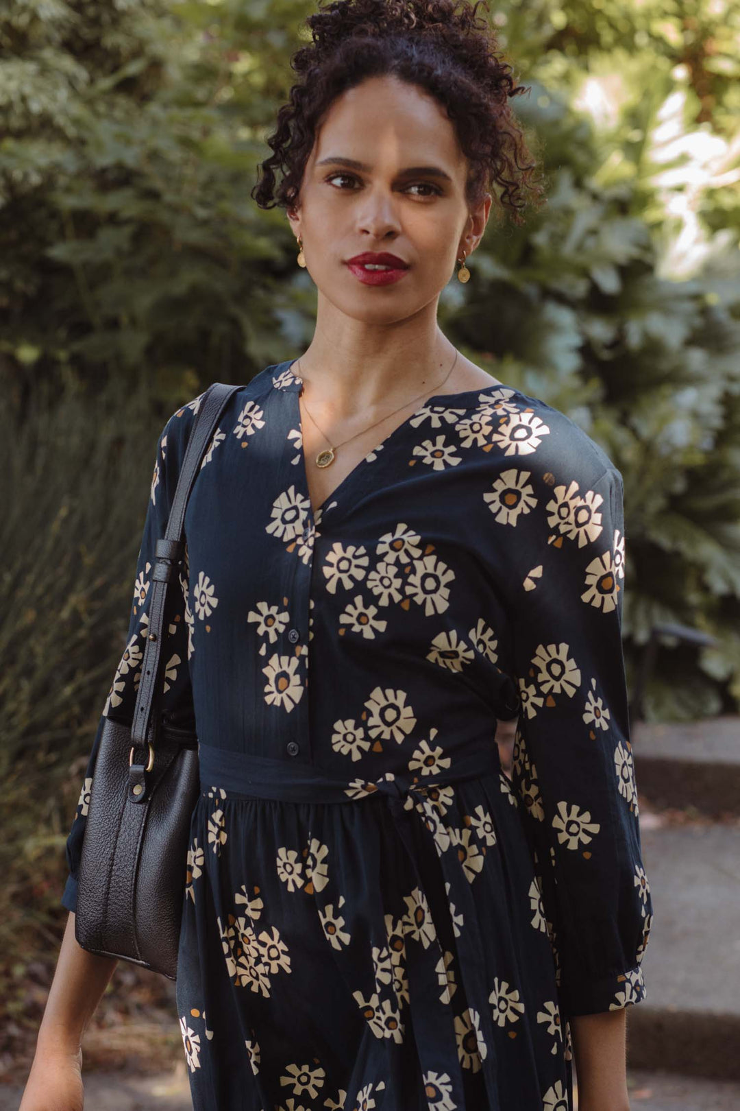 A woman with curly hair wears the Bridge & Burn Fiona Dress / Night Garden, a dark floral dress with gathered sleeves, and carries a black handbag while standing confidently outdoors amid lush greenery.