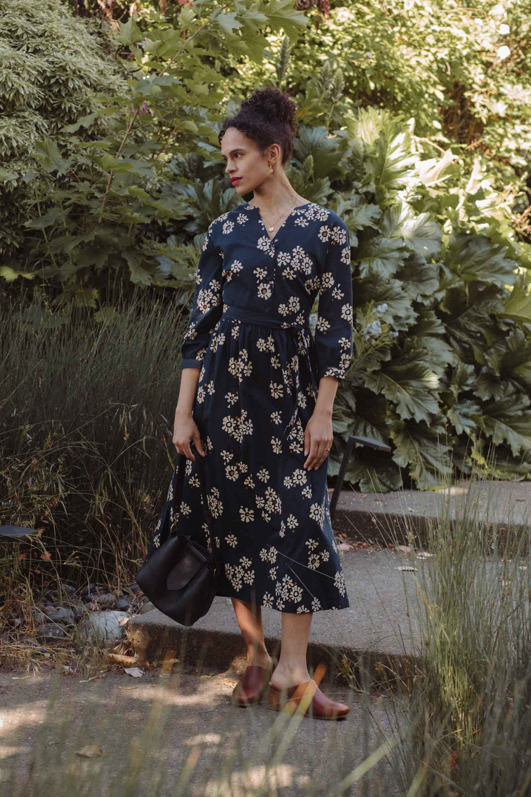 A woman stands outdoors on a stone path surrounded by greenery, wearing the Bridge & Burn Fiona Dress in Night Garden with gathered sleeves and a white floral print. She pairs it with brown flats, holds a black bag, and gazes thoughtfully to the side.