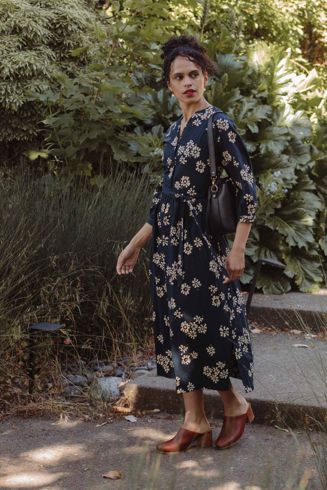 A woman wears the Bridge & Burn Fiona Dress in Night Garden as she stands on a path surrounded by greenery, carrying a black shoulder bag and wearing brown shoes, looking thoughtfully to her left.