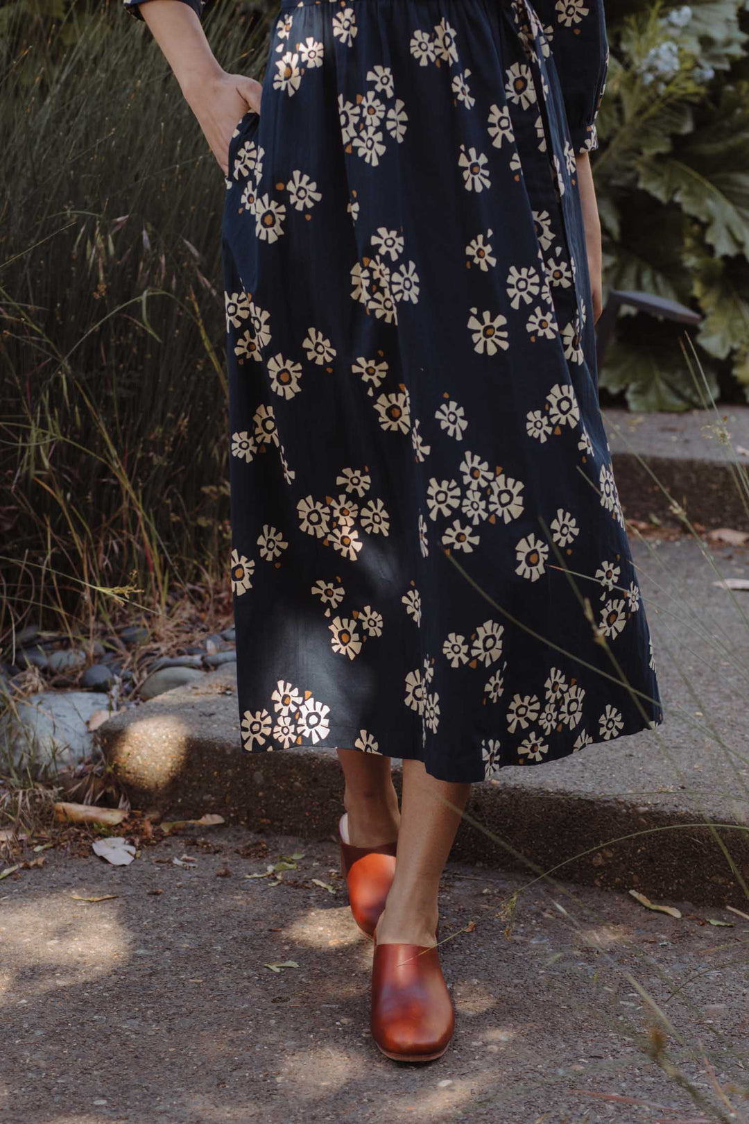 A person wearing the Fiona Dress / Night Garden by Bridge & Burn—an organic cotton navy dress with white florals—walks on a sunlit path surrounded by greenery.