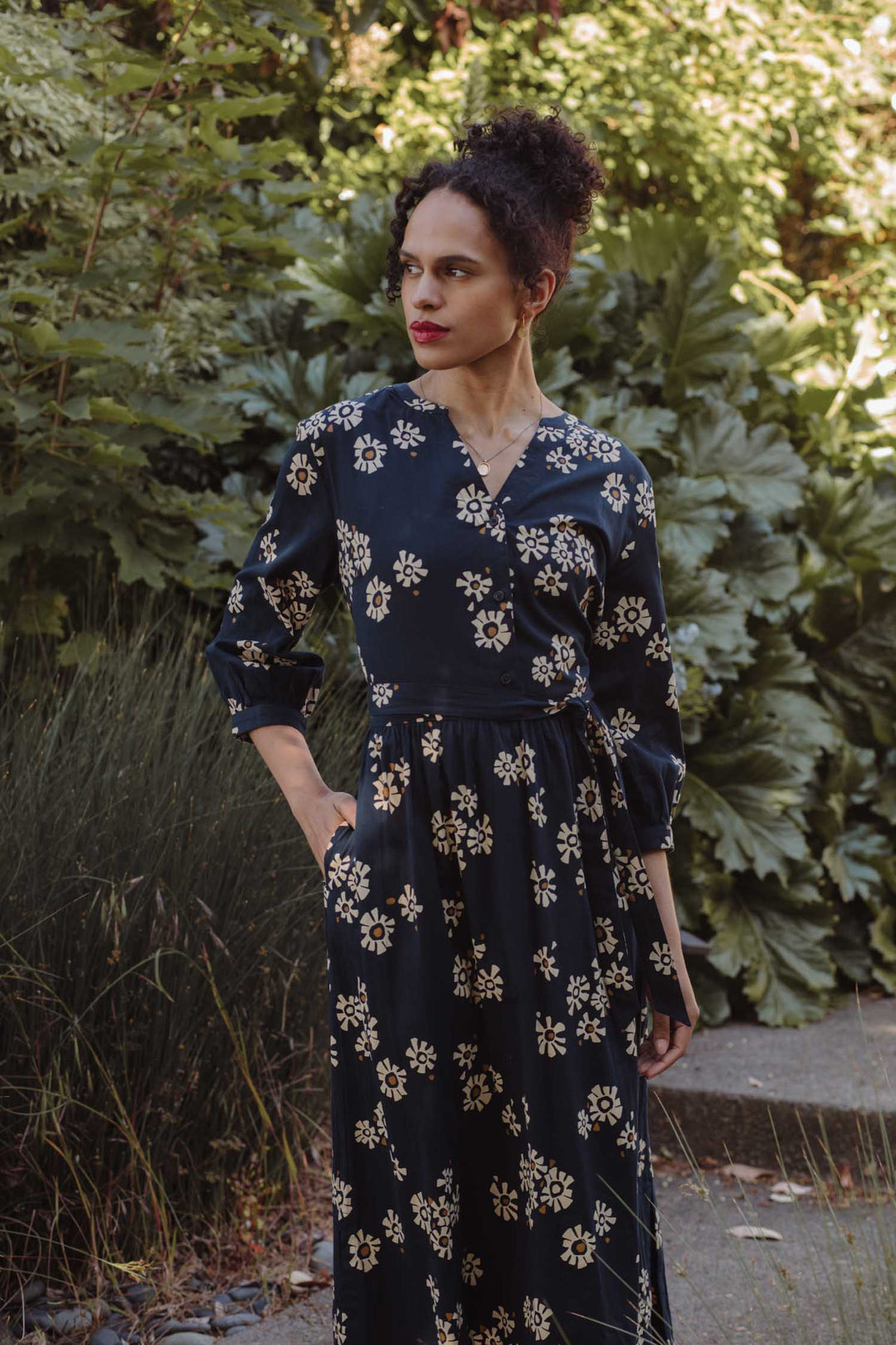A woman with curly hair in an updo stands outdoors among lush green foliage, wearing Bridge & Burn’s Fiona Dress / Night Garden—a long-sleeved, navy dress with a cream floral pattern—as she looks to the side with one hand in her pocket.