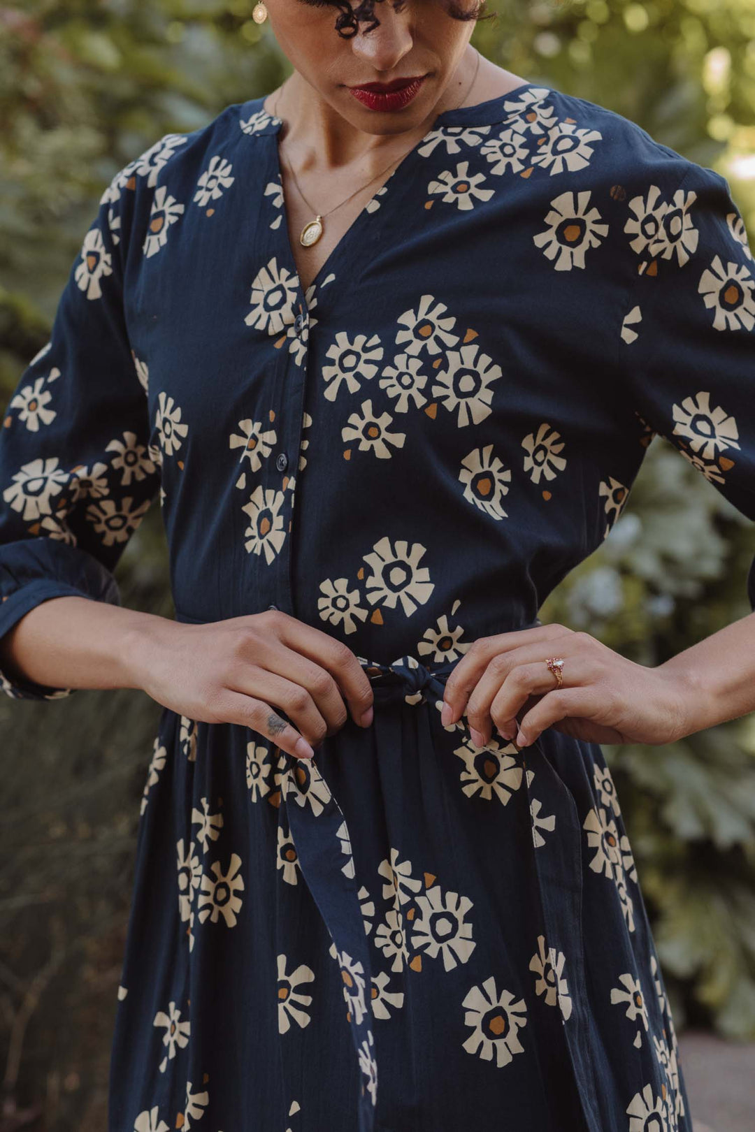 Wearing the Bridge & Burn Fiona Dress / Night Garden—a navy blue gathered sleeve dress with a white floral pattern—a person with curly hair and red lipstick adjusts the waist tie while standing outdoors among lush greenery.