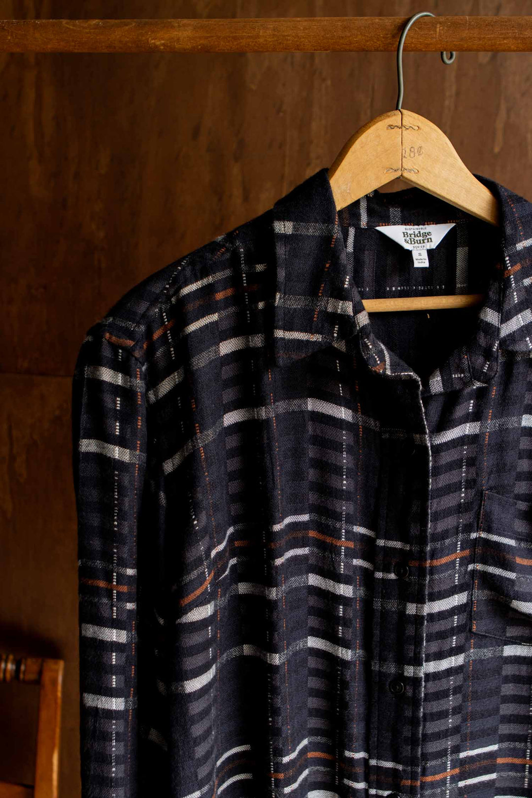 The Bridge & Burn Haley Shirt in Nightfall Stripe—a black, long-sleeve plaid shirt with white and orange stripes and a relaxed fit—hangs on a wooden hanger against a dark wooden background.