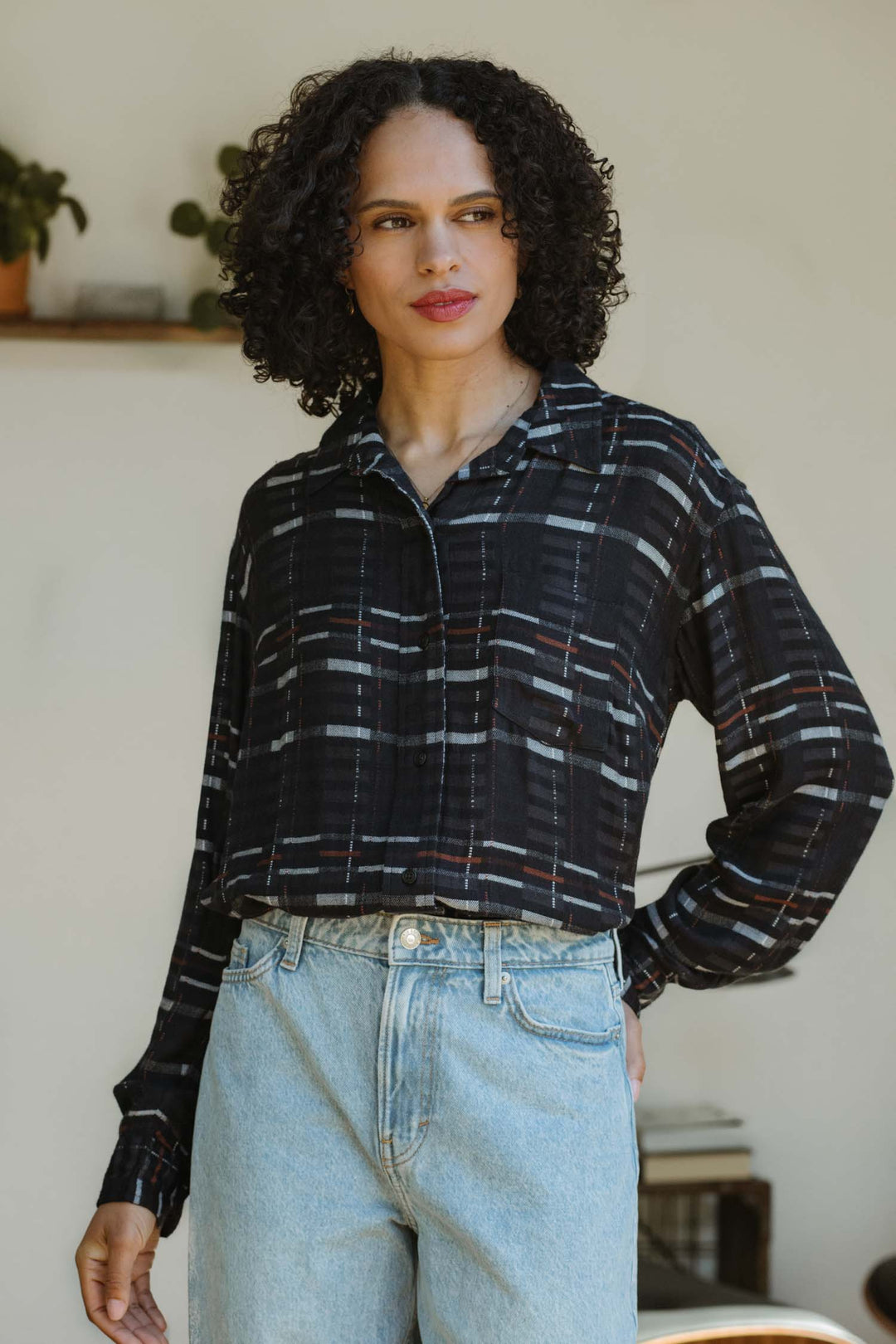 A person with curly dark hair wears a relaxed fit Bridge & Burn Haley Shirt in Nightfall Stripe and light blue jeans, standing indoors with one hand in their pocket. A plant and bookshelf are visible in the background.
