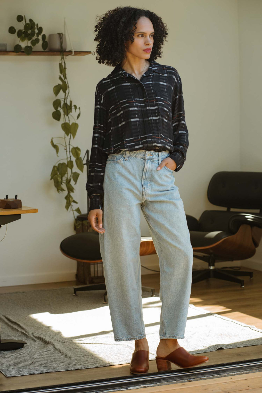 A person with curly hair wears a Bridge & Burn Haley Shirt in Nightfall Stripe, relaxed fit, paired with light blue high-waisted jeans and brown shoes, standing in a sunlit room with modern furniture and a wall-mounted plant.