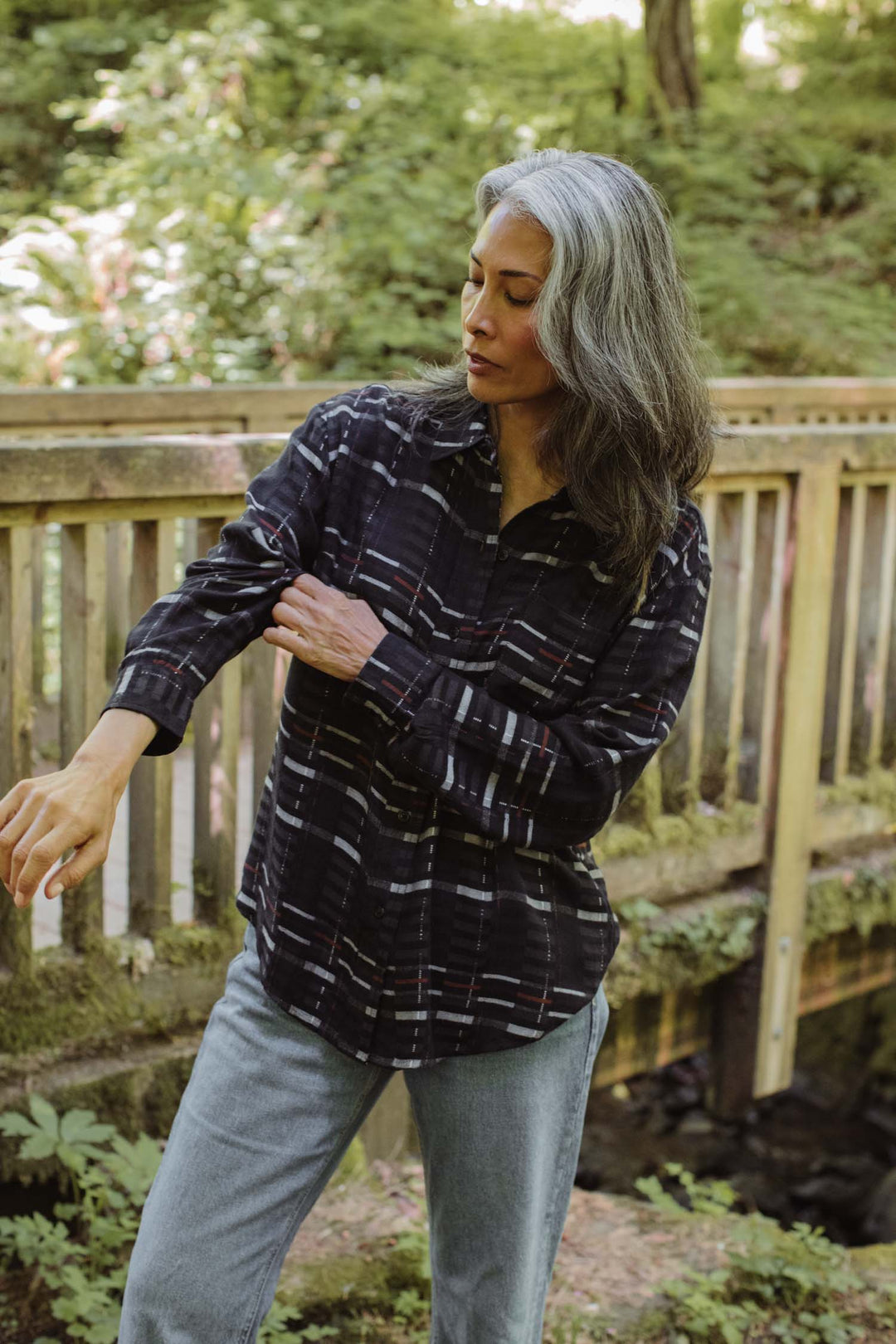 A woman with long gray hair stands outdoors near a wooden railing, rolling up the sleeves of her Bridge & Burn Haley Shirt in Nightfall Stripe, paired with jeans and surrounded by green foliage.