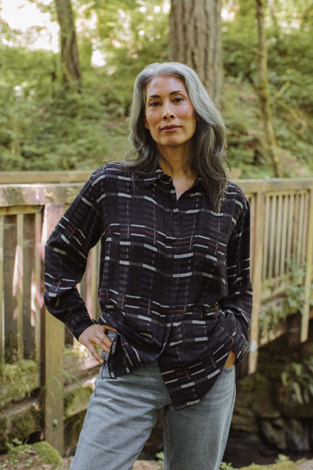 A woman with long gray hair stands outdoors by a wooden railing, surrounded by lush greenery, wearing the relaxed fit Haley Shirt in Nightfall Stripe from Bridge & Burn with light jeans and one hand on her hip.