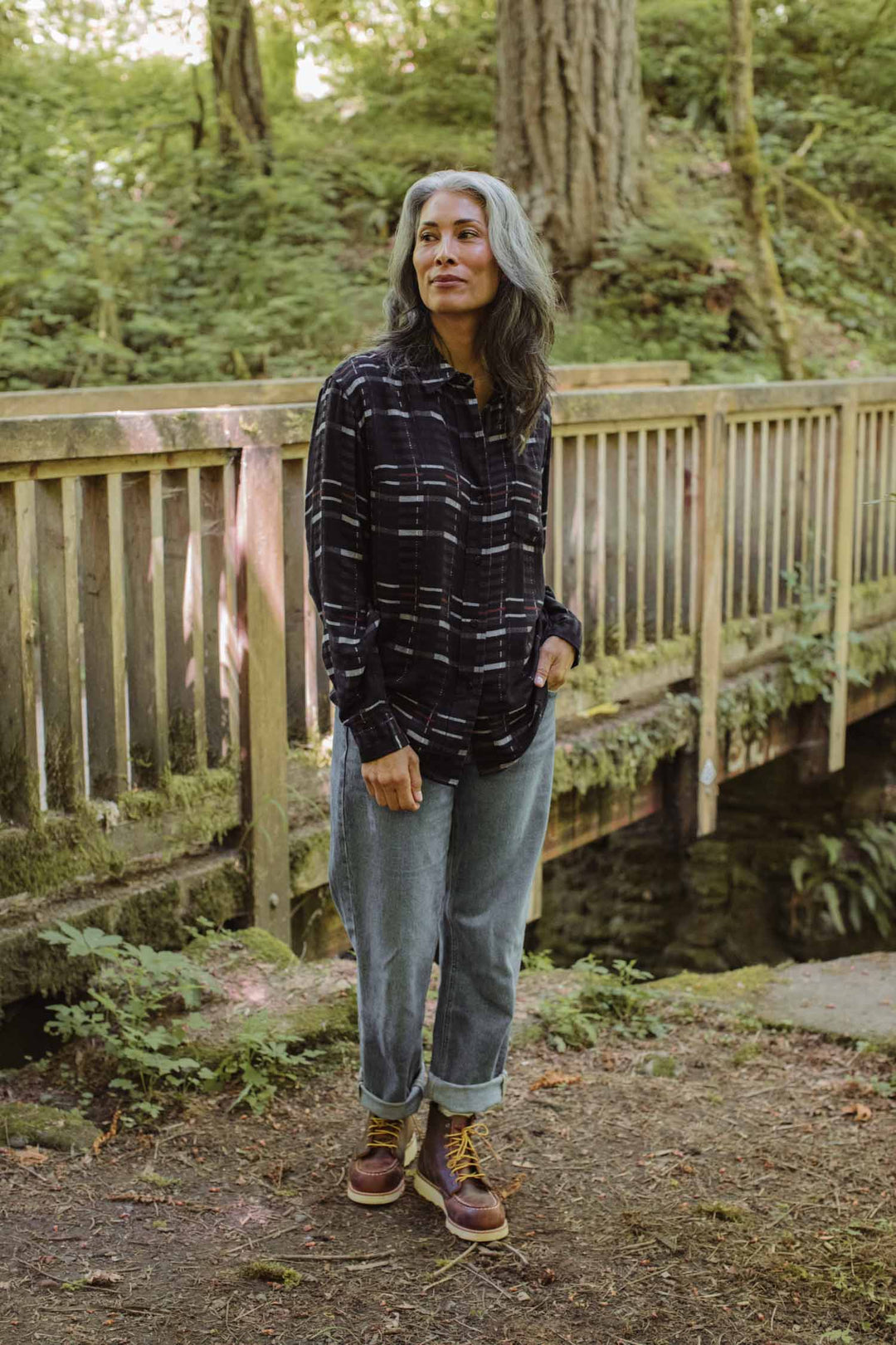 A person with long gray hair stands outdoors by a wooden railing, wearing the Bridge & Burn Haley Shirt in Nightfall Stripe, rolled-up jeans, and brown boots, surrounded by lush green trees.