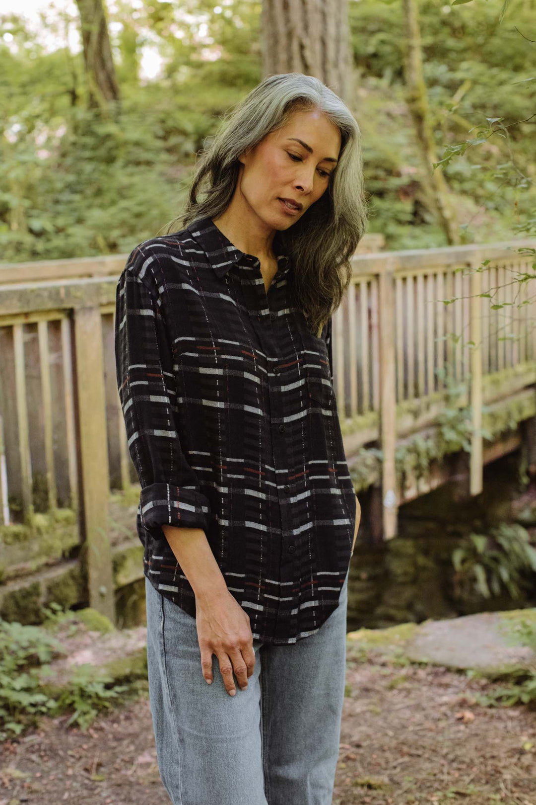 A woman with long gray hair, dressed in the Bridge & Burn Haley Shirt in Nightfall Stripe and jeans, stands outdoors by a wooden bridge surrounded by greenery, gazing down thoughtfully.