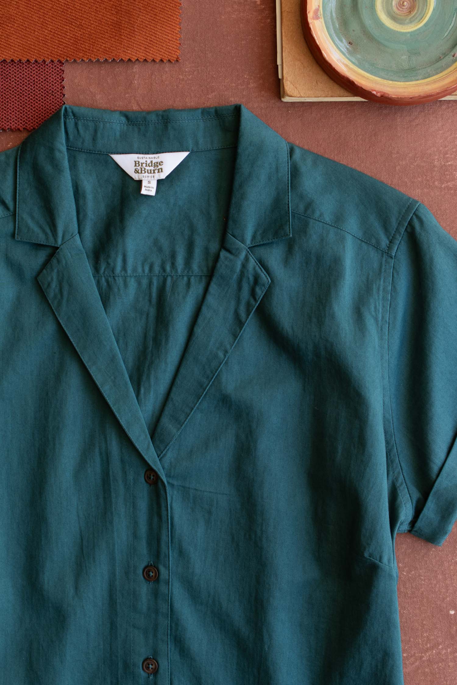 The Bridge & Burn Innes Shirt in Deep Sea, a teal short-sleeve button-up with a notched collar and relaxed fit, is laid flat on a brown surface beside fabric swatches and a round ceramic plate featuring a spiral pattern.