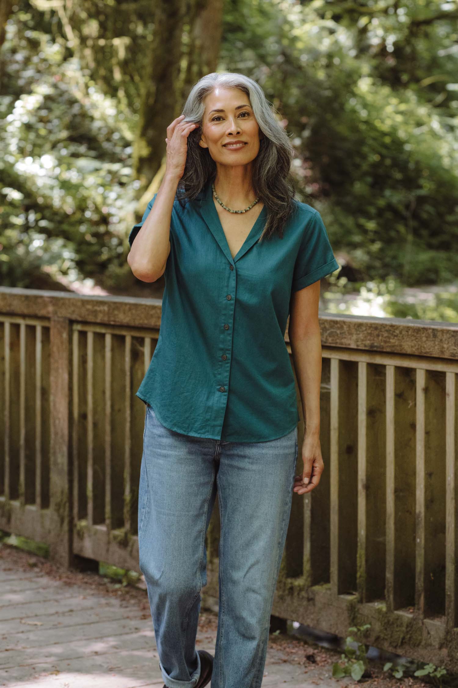 A woman with long gray hair wears the Bridge & Burn Innes Shirt in Deep Sea and jeans, standing on a wooden bridge in a lush green setting. She touches her hair and smiles gently at the camera.