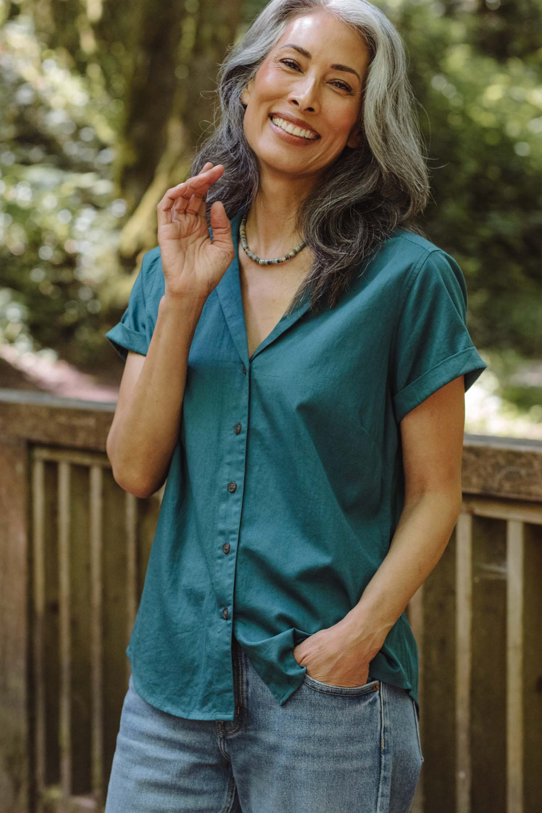 A smiling woman with long gray hair wears the Bridge & Burn Innes Shirt in Deep Sea and jeans as she stands outdoors by a wooden railing, green trees in the background.