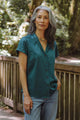 A woman with long gray hair, dressed in the Bridge & Burn Innes Shirt in Deep Sea and jeans, stands outdoors by a wooden railing surrounded by trees and greenery.