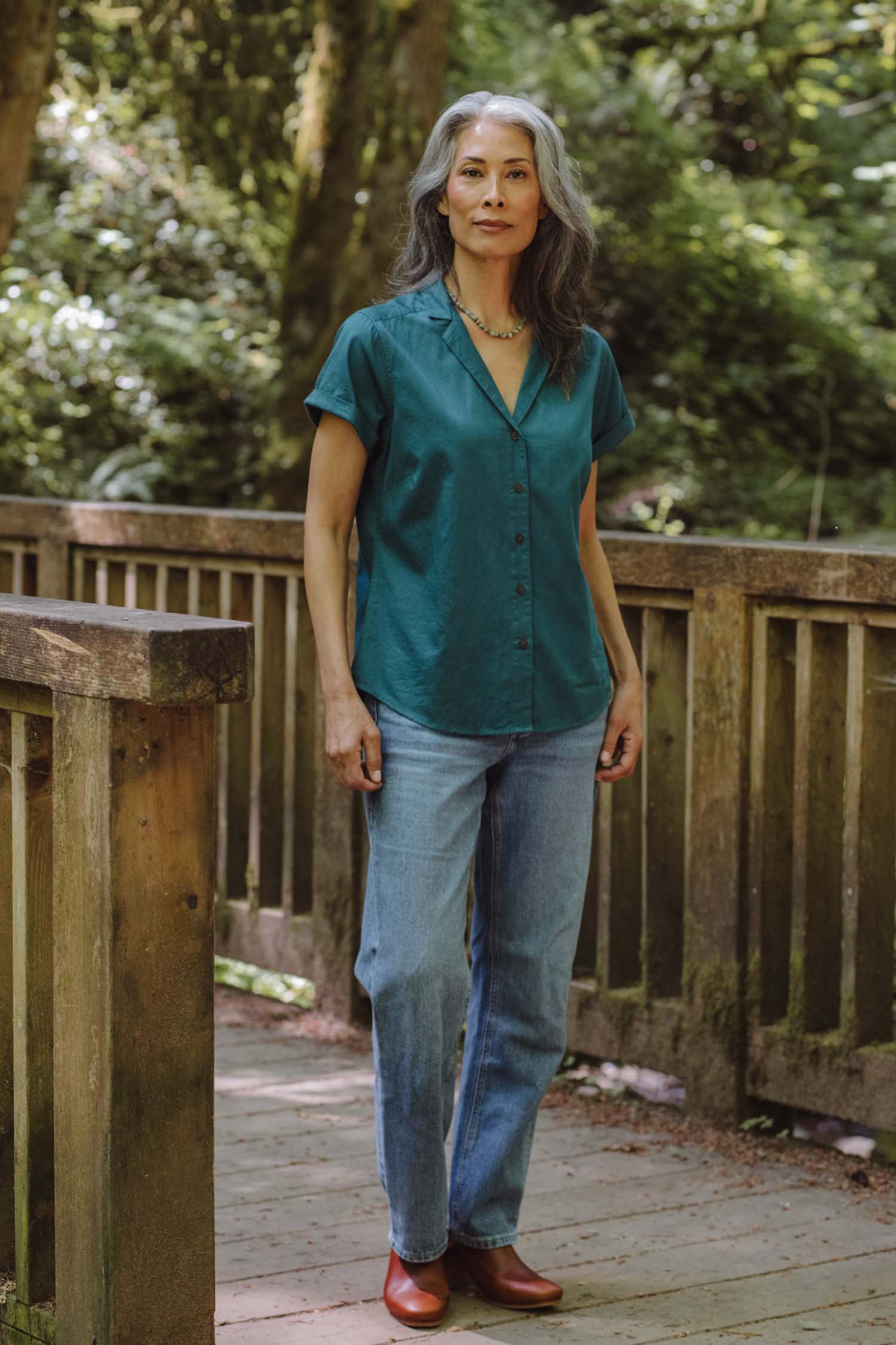 A woman with long gray hair stands on a wooden bridge in a forest, wearing the Bridge & Burn Innes Shirt in Deep Sea, blue jeans, and red shoes. Lush green foliage surrounds her in the background.