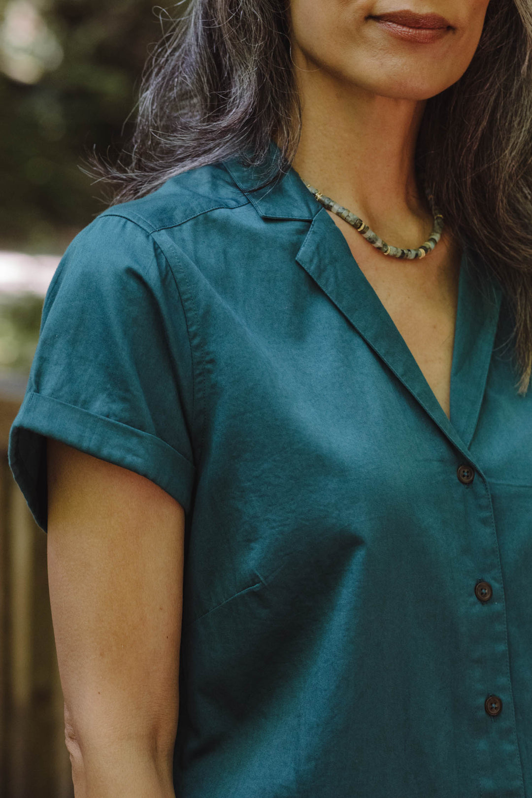 A woman with long dark hair wears the Bridge & Burn Innes Shirt in Deep Sea, styled with a beaded necklace. Standing outdoors, she is shown from shoulders to mid-arm in this cropped image.