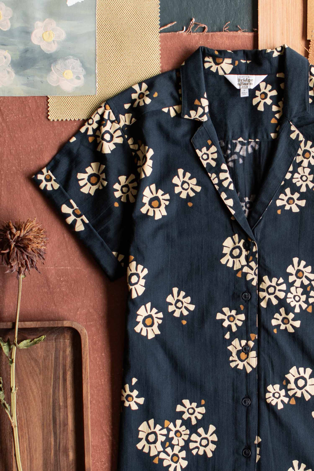 The Innes Shirt / Night Garden by Bridge & Burn is a relaxed-fit, short sleeve shirt in navy with a beige floral pattern, crafted from organic cotton for comfort. Shown on a brown surface beside dried flowers, wood tray, swatches, and art.