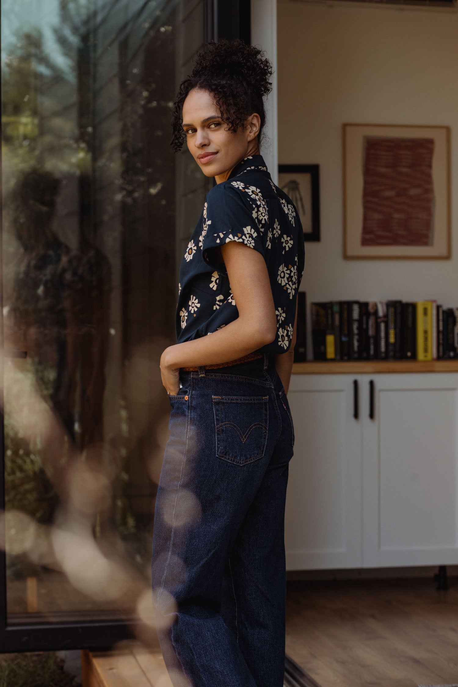 A person with curly hair, dressed in the Bridge & Burn Innes Shirt in Night Garden and blue jeans, stands indoors by an open glass door, glancing over their shoulder. Books and framed art sit on a white cabinet behind them.