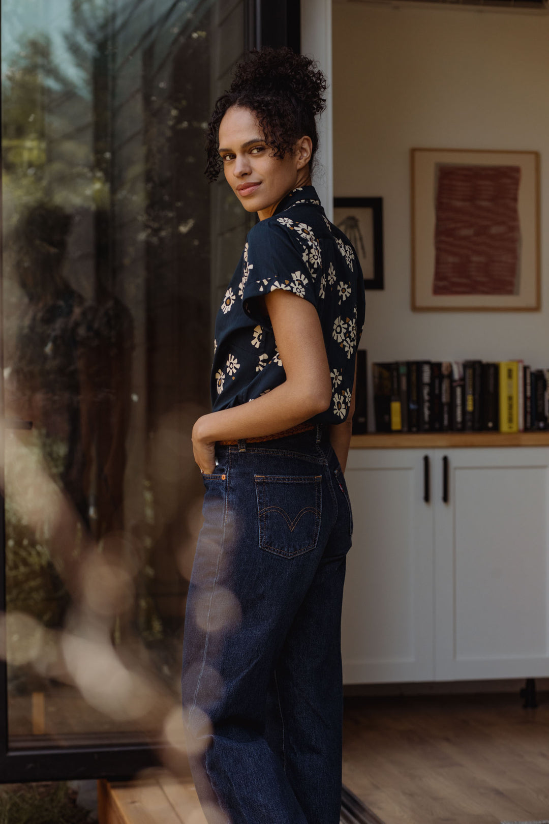 A person with curly hair, dressed in the Bridge & Burn Innes Shirt in Night Garden and blue jeans, stands indoors by an open glass door, glancing over their shoulder. Books and framed art sit on a white cabinet behind them.