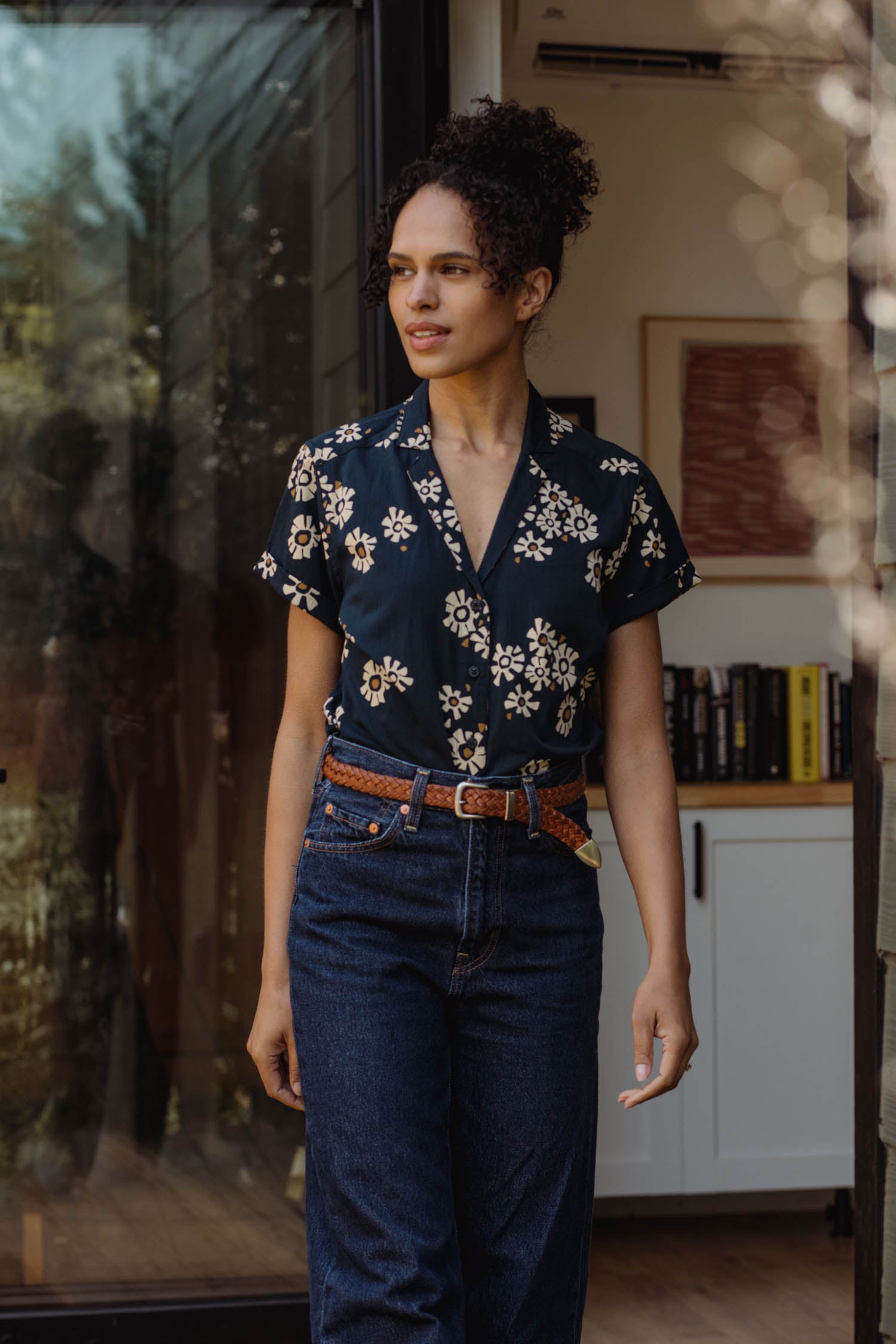 A woman wears the Bridge & Burn Innes Shirt in Night Garden, tucked into high-waisted jeans with a brown belt, standing in a sunlit doorway with books and artwork visible inside the room behind her.
