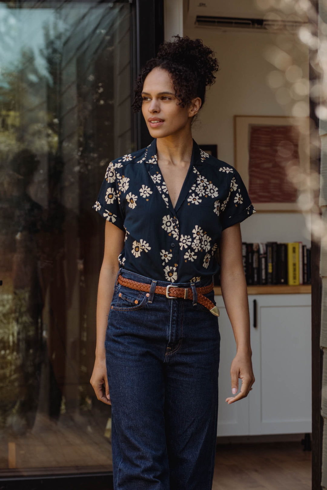 A woman wears the Bridge & Burn Innes Shirt in Night Garden, tucked into high-waisted jeans with a brown belt, standing in a sunlit doorway with books and artwork visible inside the room behind her.