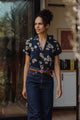 A woman wears the Bridge & Burn Innes Shirt in Night Garden, tucked into high-waisted jeans with a brown belt, standing in a sunlit doorway with books and artwork visible inside the room behind her.