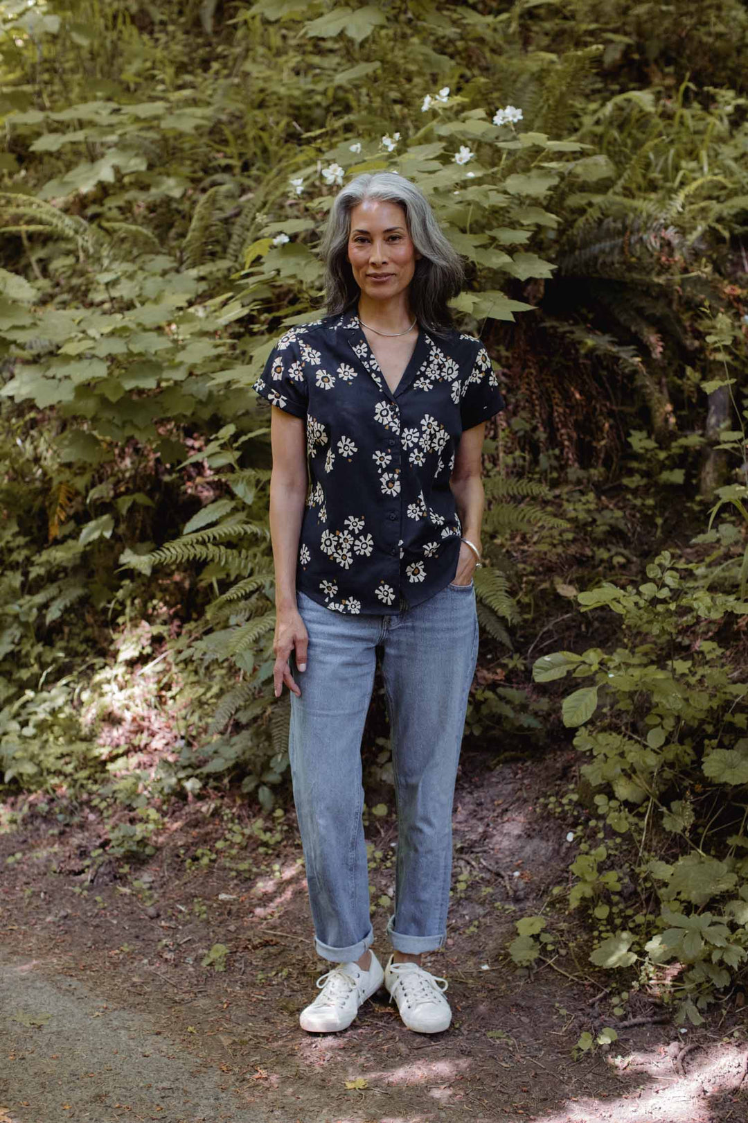 A woman with gray hair stands on a forest path, wearing the Bridge & Burn Innes Shirt in Night Garden, paired with cuffed jeans and white sneakers. Green foliage and leafy plants surround her.