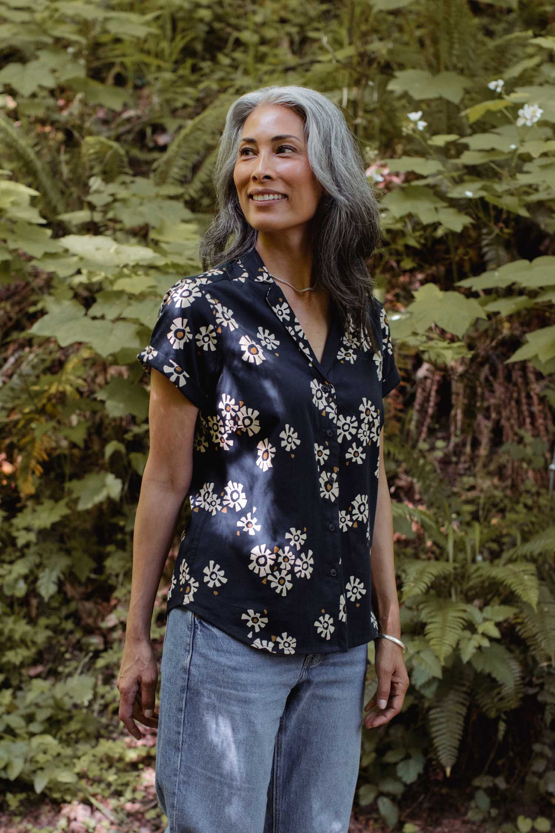 A woman with long gray hair smiles outdoors, wearing the Bridge & Burn Innes Shirt in the Night Garden print and light jeans, with green foliage and white flowers in the background.