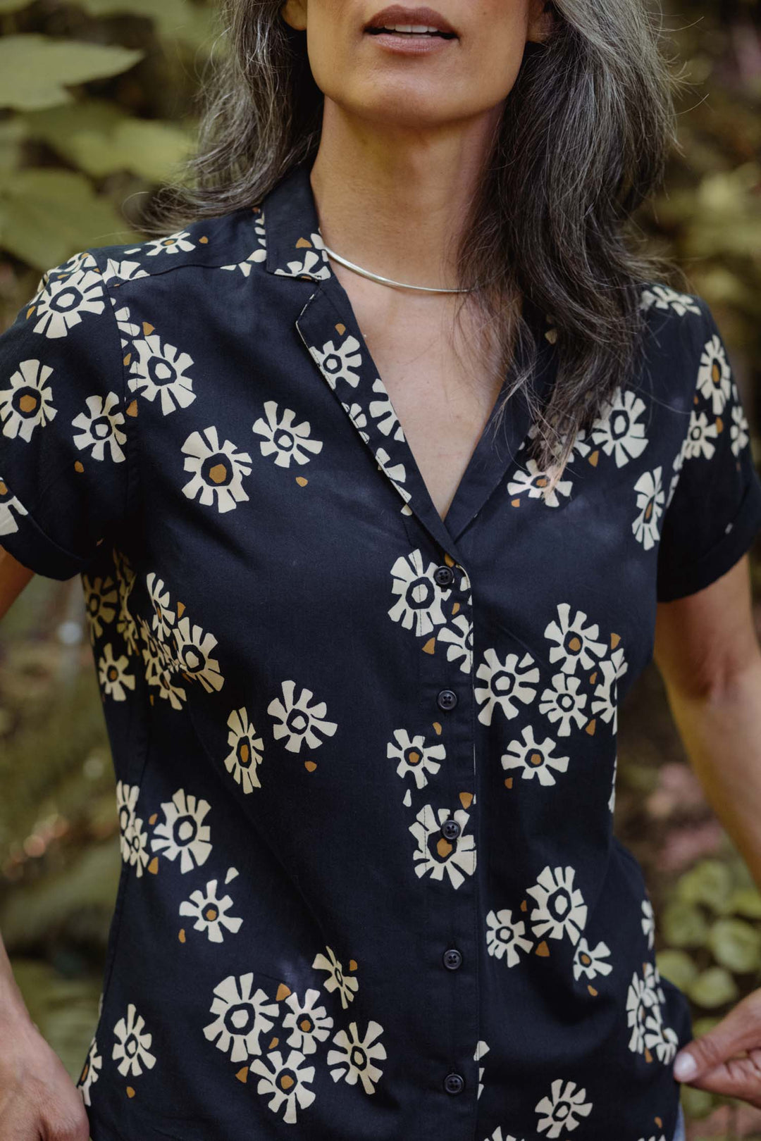 A person with long, gray hair wears the Bridge & Burn Innes Shirt in Night Garden—a relaxed fit organic cotton short sleeve button-up with cream floral pattern—and a simple silver necklace, standing outdoors among greenery.