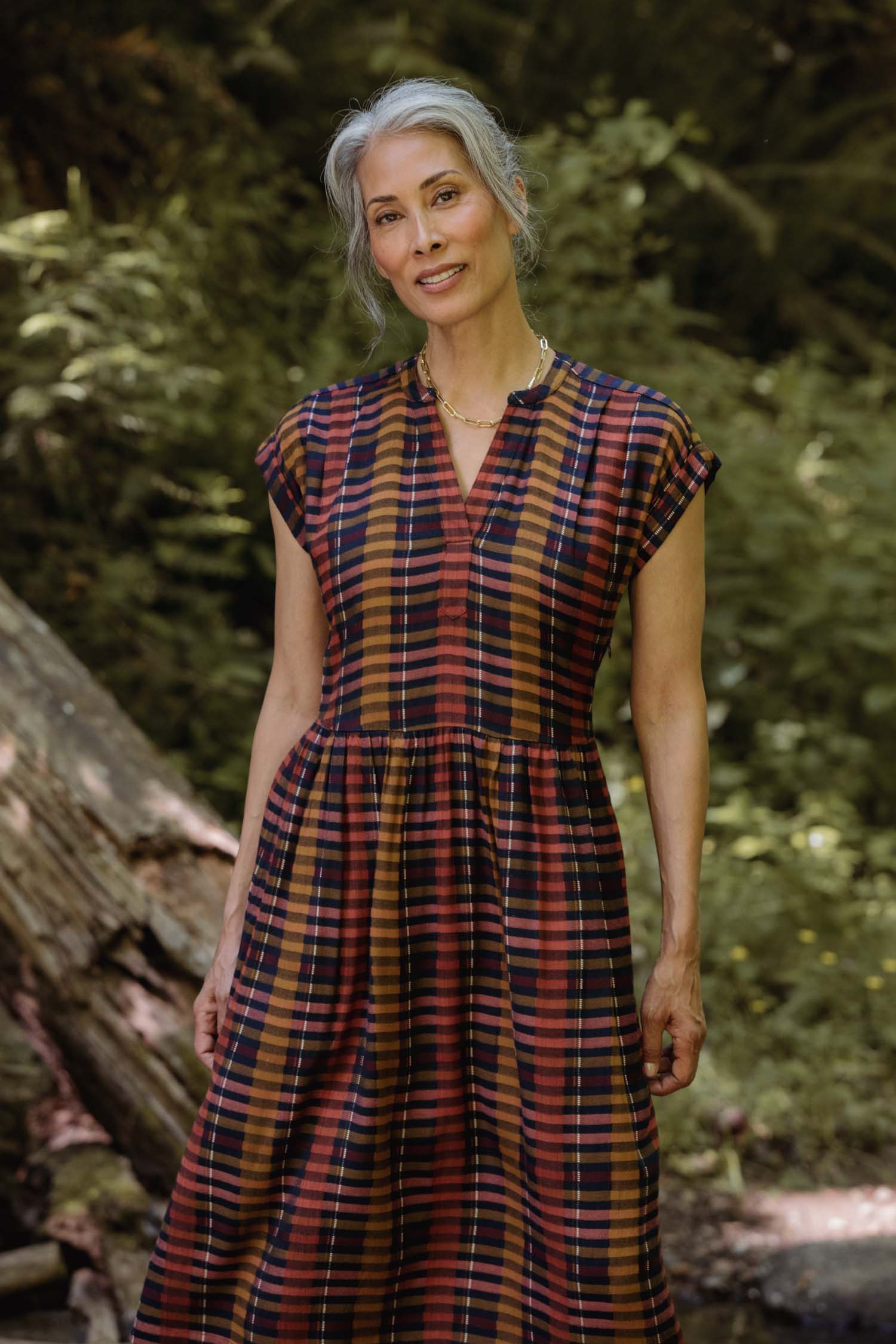 A woman with gray hair stands outdoors in a forest, softly smiling as sunlight filters through the trees. She wears the Bridge & Burn Ira Dress in Alpenglow Stripe, the short-sleeved pullover bright against the lush greenery.