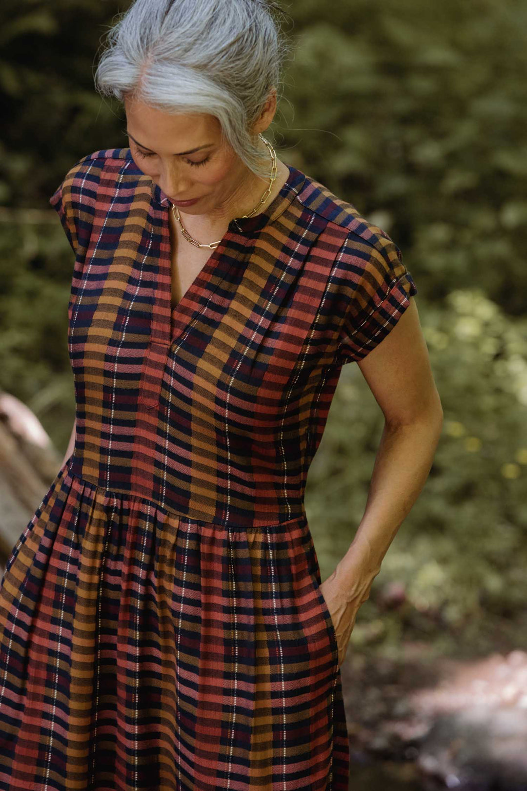 A person with gray hair pulled back looks down while wearing the Bridge & Burn Ira Dress in Alpenglow Stripe—an eco-friendly viscose, short-sleeved, plaid pullover in red, orange, yellow, and black—standing outdoors against a blurred green background.