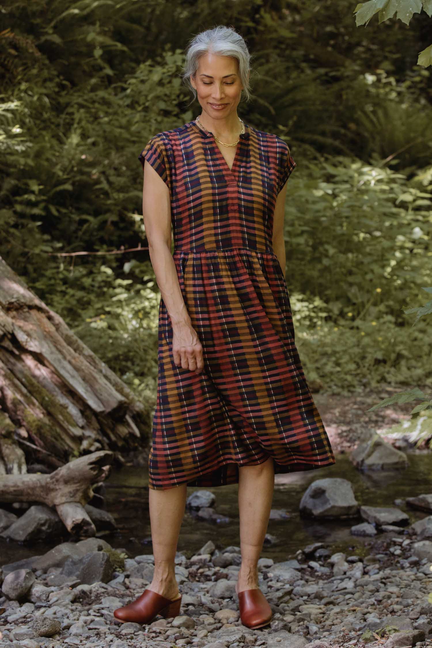 A woman with gray hair stands outdoors on rocky ground, surrounded by greenery and trees, wearing the Bridge & Burn Ira Dress in Alpenglow Stripe with brown shoes.