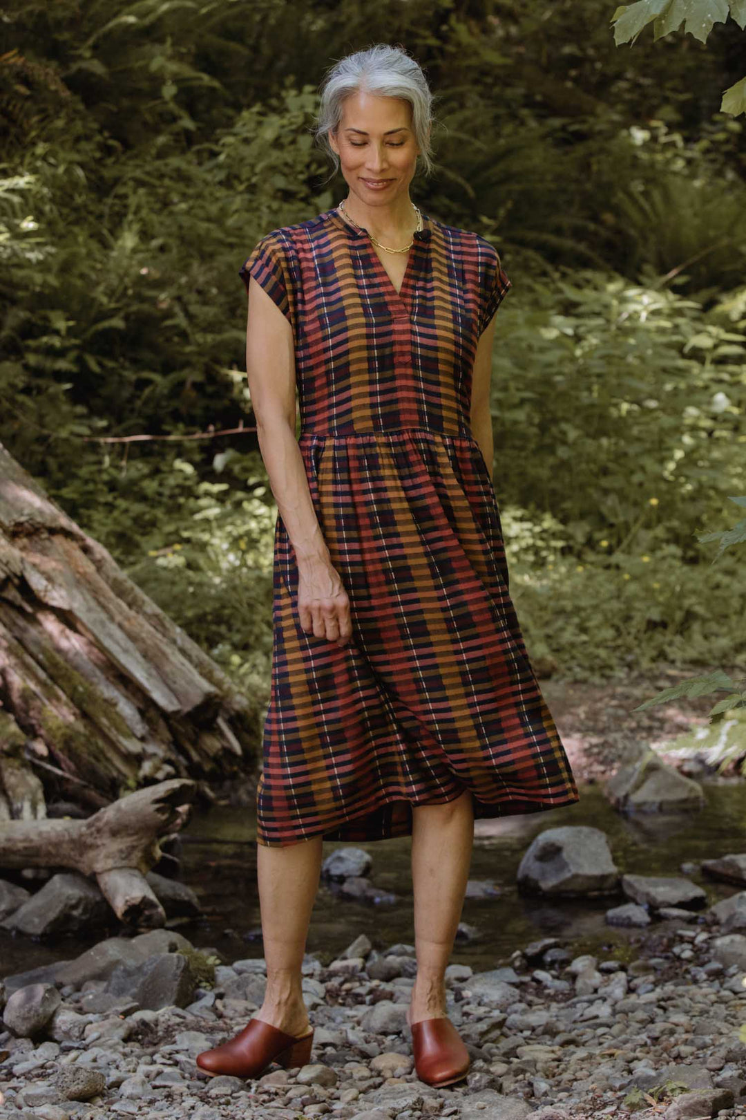 A woman with gray hair stands outdoors on rocky ground, surrounded by greenery and trees, wearing the Bridge & Burn Ira Dress in Alpenglow Stripe with brown shoes.