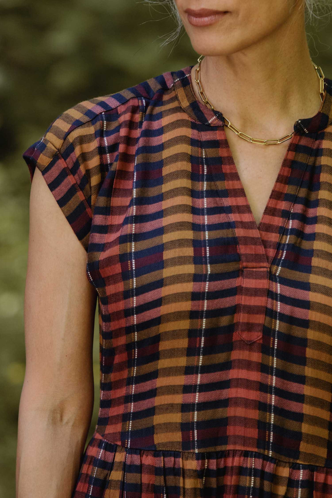 A woman stands outdoors, partially visible from shoulders to above the waist, wearing a gold chain necklace and the Bridge & Burn Ira Dress in Alpenglow Stripe—a short-sleeved, plaid pullover in red, brown, and navy tones.