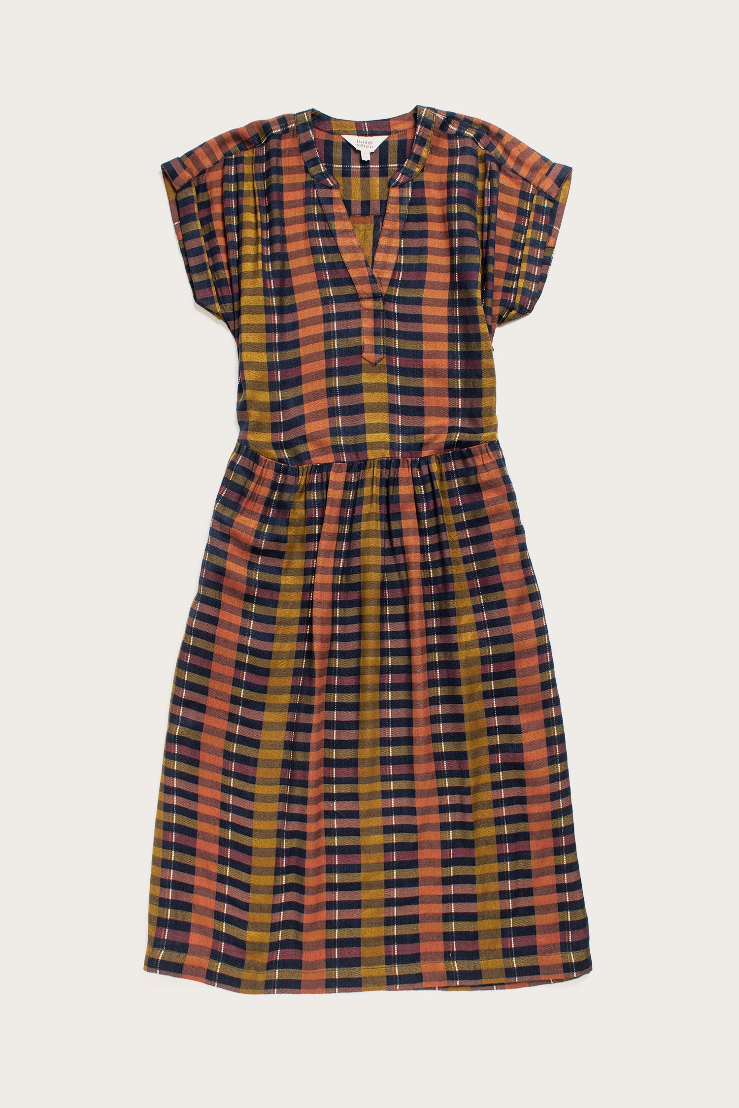 The Ira Dress by Bridge & Burn features knee-length, short dolman sleeves, a v-neck, Alpenglow Stripe pattern in mustard yellow, rust orange, navy blue, and black. Made from eco-friendly viscose with a gathered waist and relaxed fit.