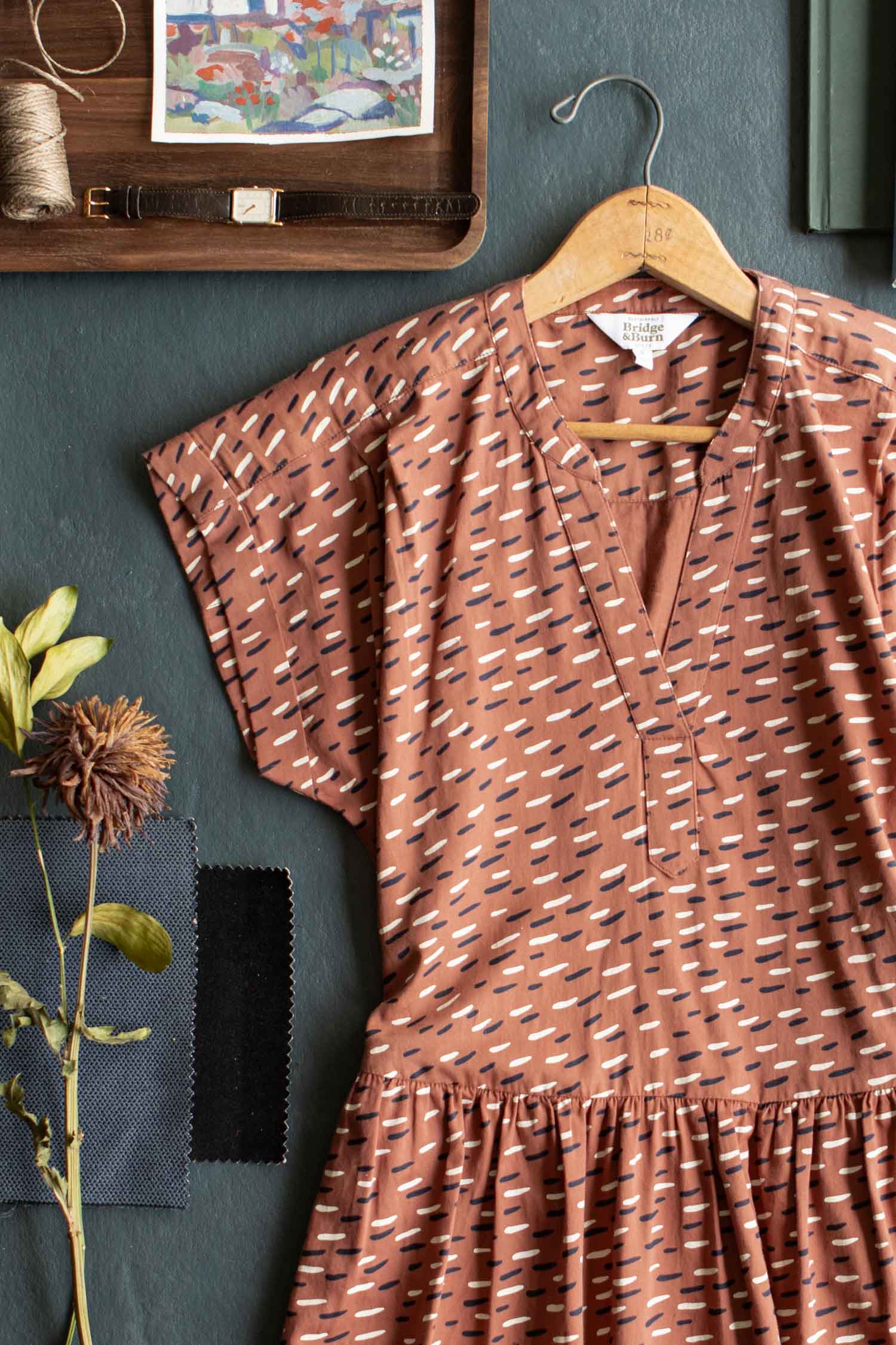 The Ira Dress / Desert Rain by Bridge & Burn, a brown organic cotton pullover with a scatter print, hangs on a wooden hanger against a dark background, surrounded by fabric swatches, dried flowers, thread, and sewing tools.