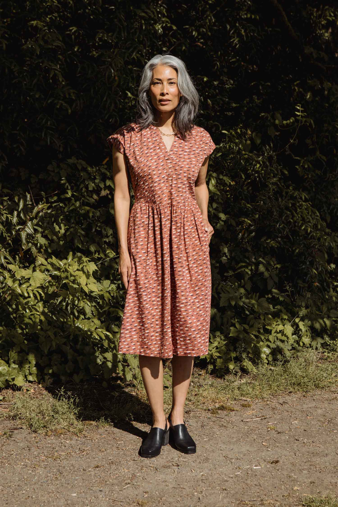 A woman with gray hair stands outdoors on a dirt path, wearing the Bridge & Burn Ira Dress in Desert Rain. The knee-length organic cotton dress features short sleeves and pockets. She pairs it with black shoes amidst lush greenery.