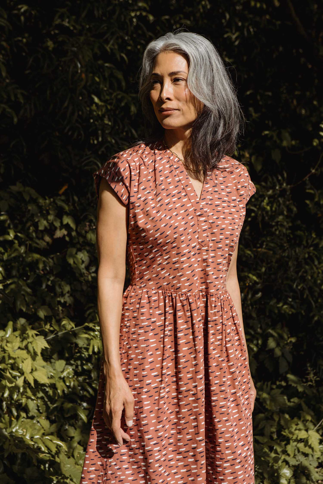 A person with long, wavy gray hair stands outdoors in lush greenery, wearing the Bridge & Burn Ira Dress in Desert Rain—an organic cotton, rust-orange patterned dress with short sleeves.