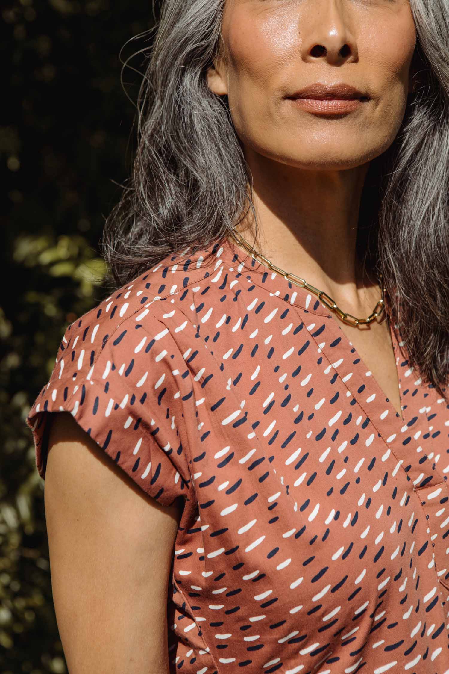 A woman with long gray hair, wearing a gold chain and the Bridge & Burn Ira Dress in Desert Rain—a rust-colored organic cotton piece with white and navy dash patterns and dolman sleeves—stands outside in sunlight, her upper face visible.