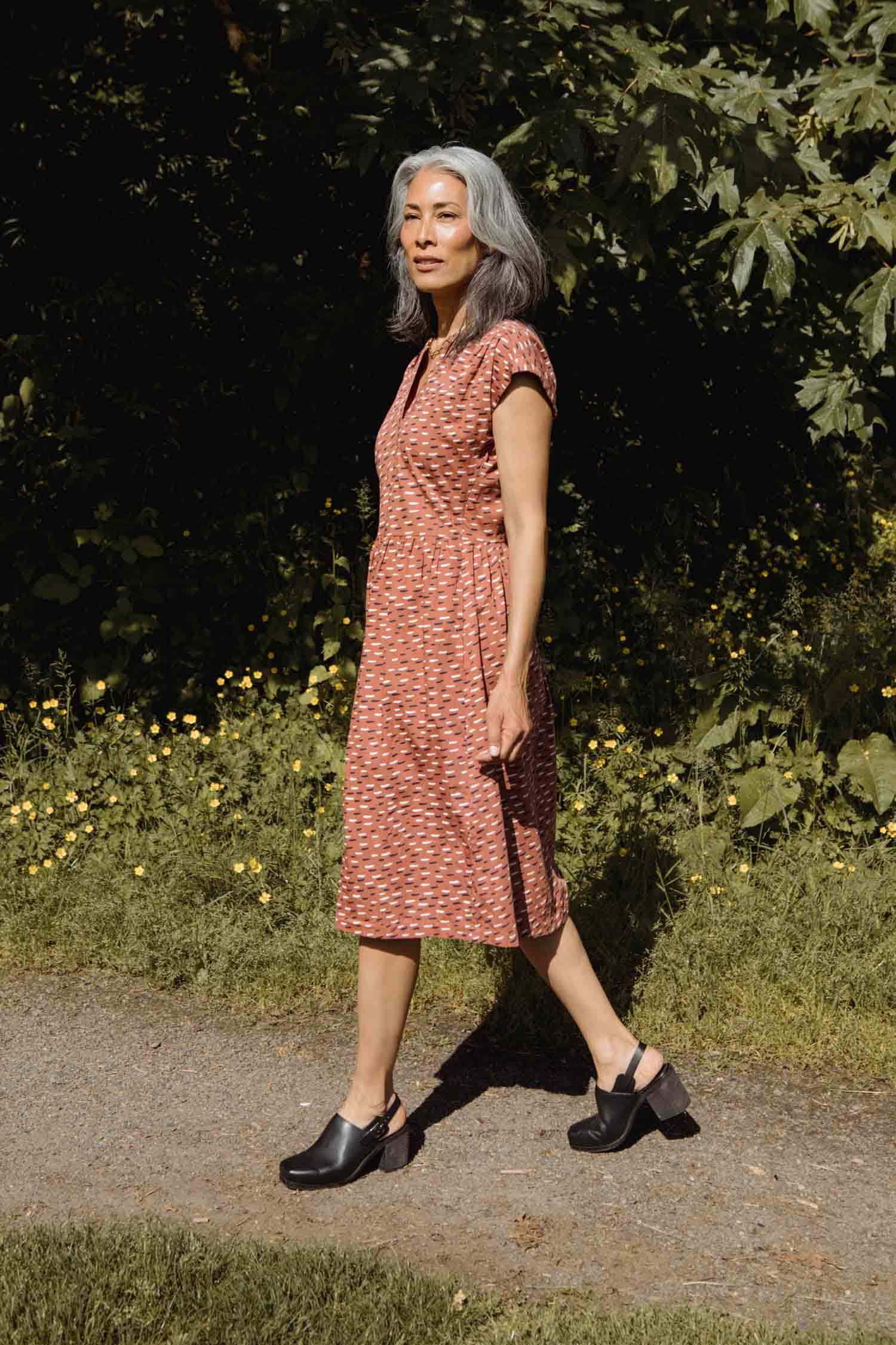 A woman with long gray hair walks along a sunlit path, surrounded by greenery and yellow wildflowers, wearing the Bridge & Burn Ira Dress in Desert Rain with black shoes.
